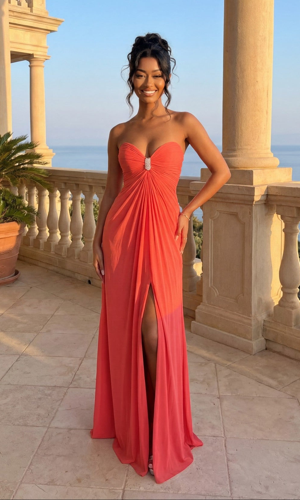 A woman stands on a stone terrace with ocean views, wearing La Femme Dress 21233 in Pink Grapefruit, featuring a strapless bodice and front slit.